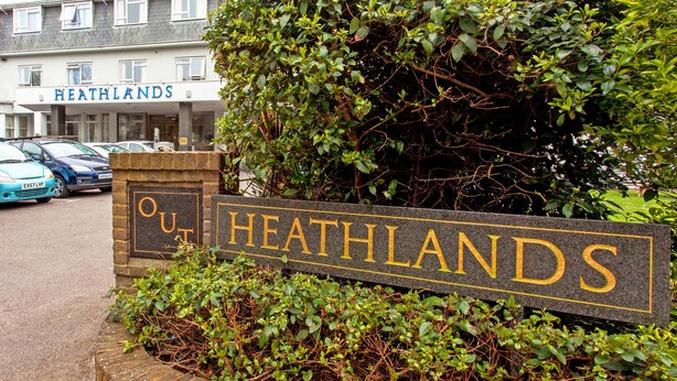 Segnaletica per 'HEATHLANDS' con base in pietra e scritte dorate, circondata dal verde.