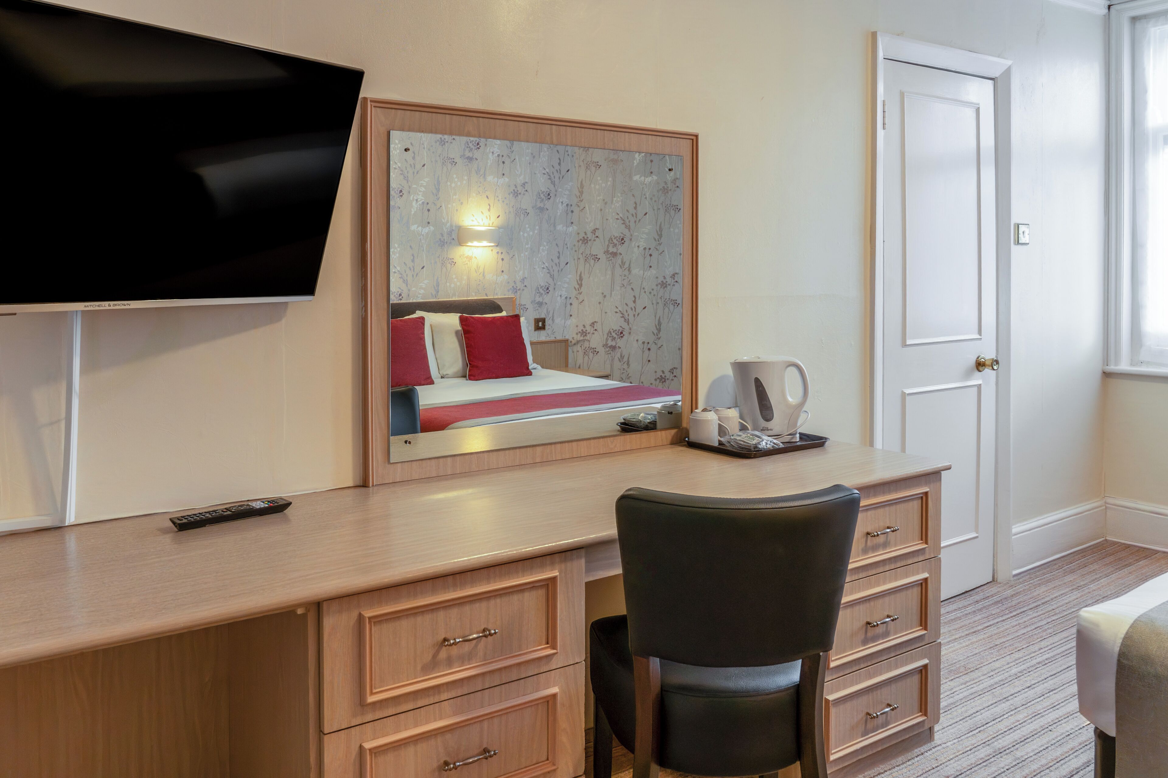A hotel room with a bed, a desk with a chair, a television, a mirror, and a door.