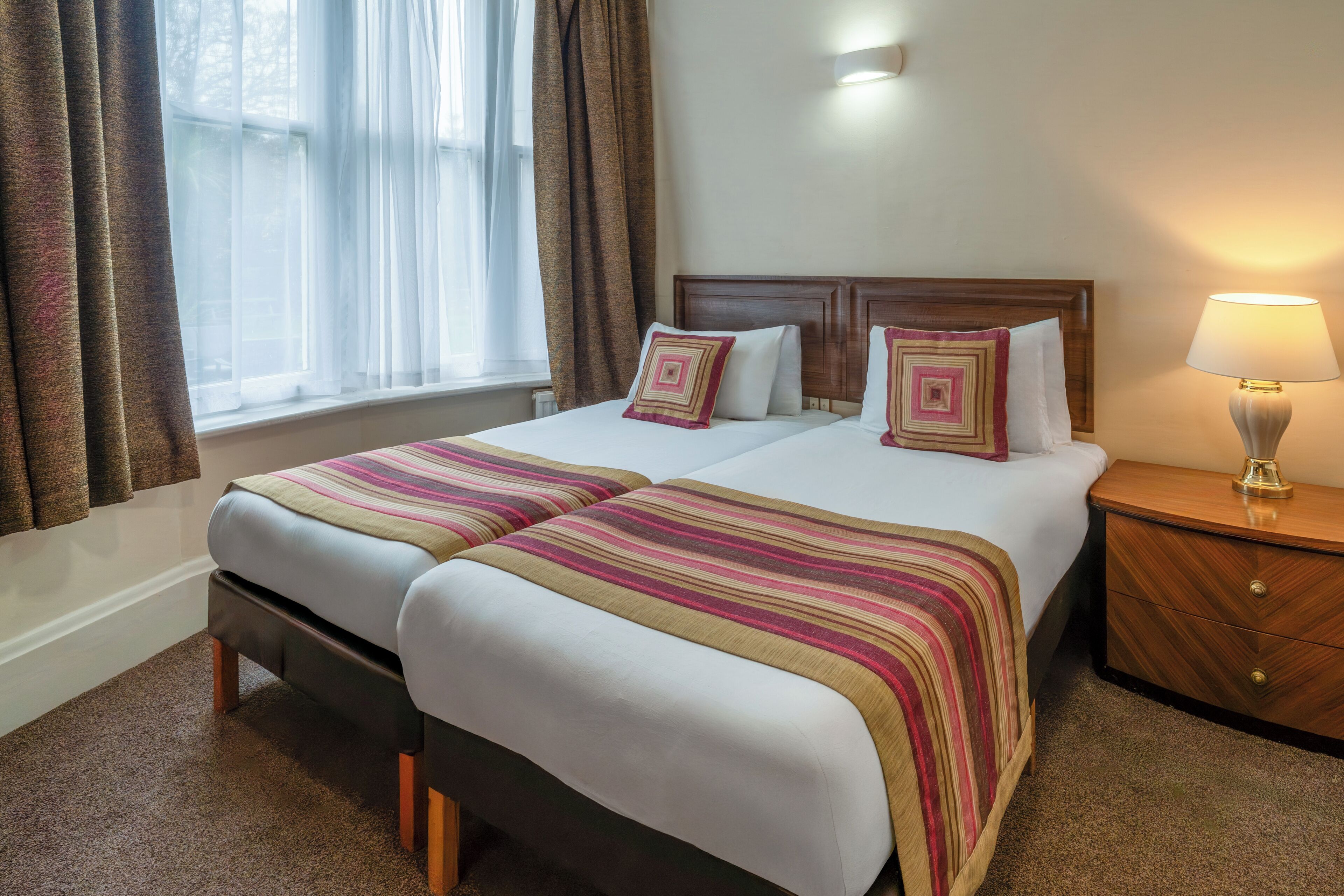 A double bed with two pillows, a bedside table with a lamp, and a window with curtains.