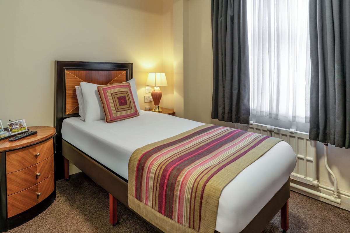 A hotel room with a bed, a nightstand, a lamp, a window with curtains, and a radiator.