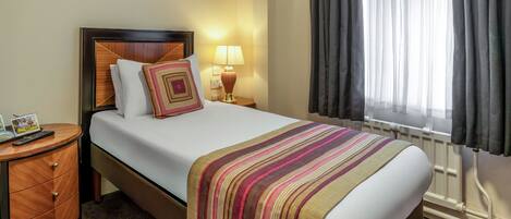 Una habitación de hotel con cama, mesita de noche, lámpara, ventana con cortinas y radiador.