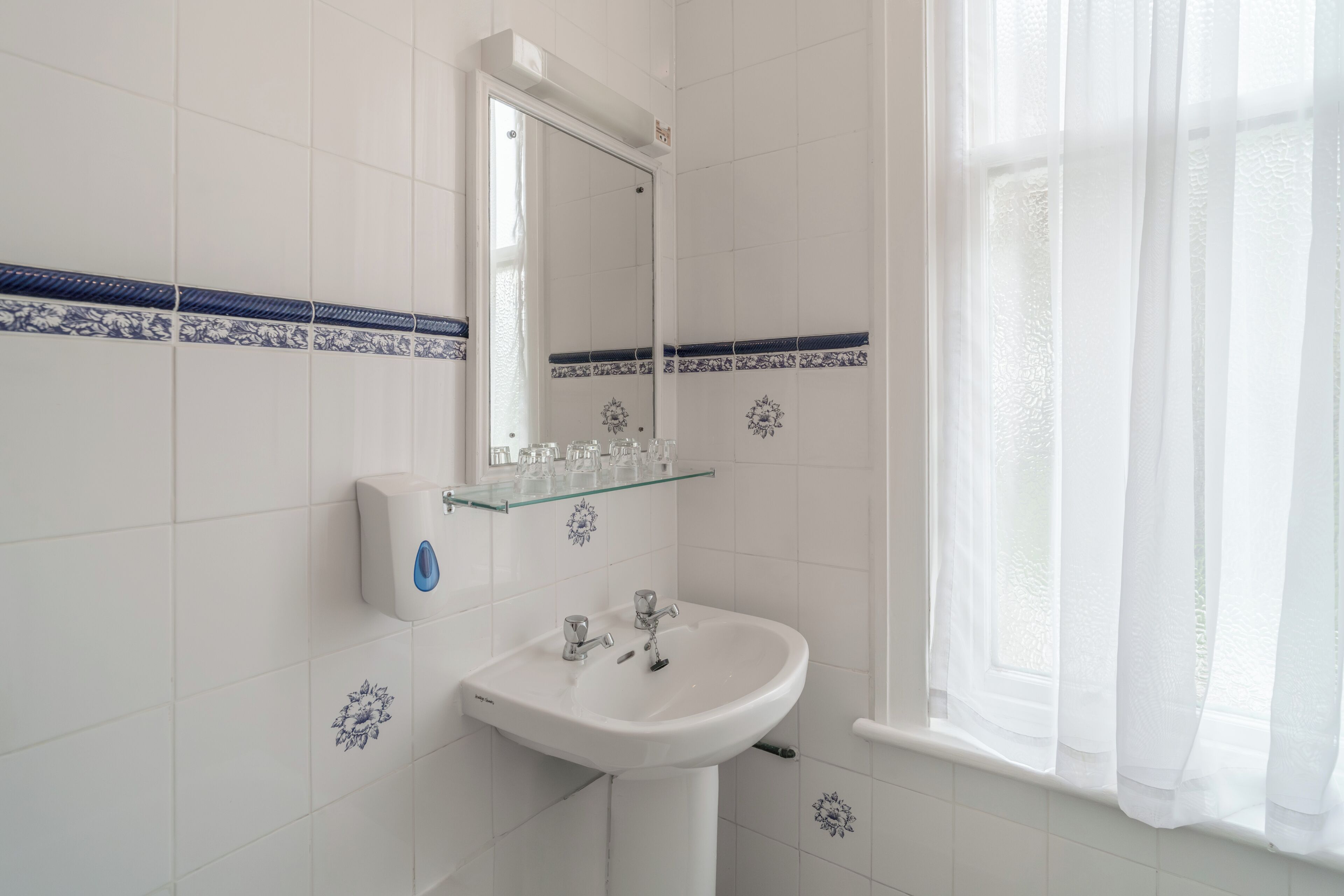 A bathroom with a white sink, a glass shelf, a mirror, and a window with sheer curtains.