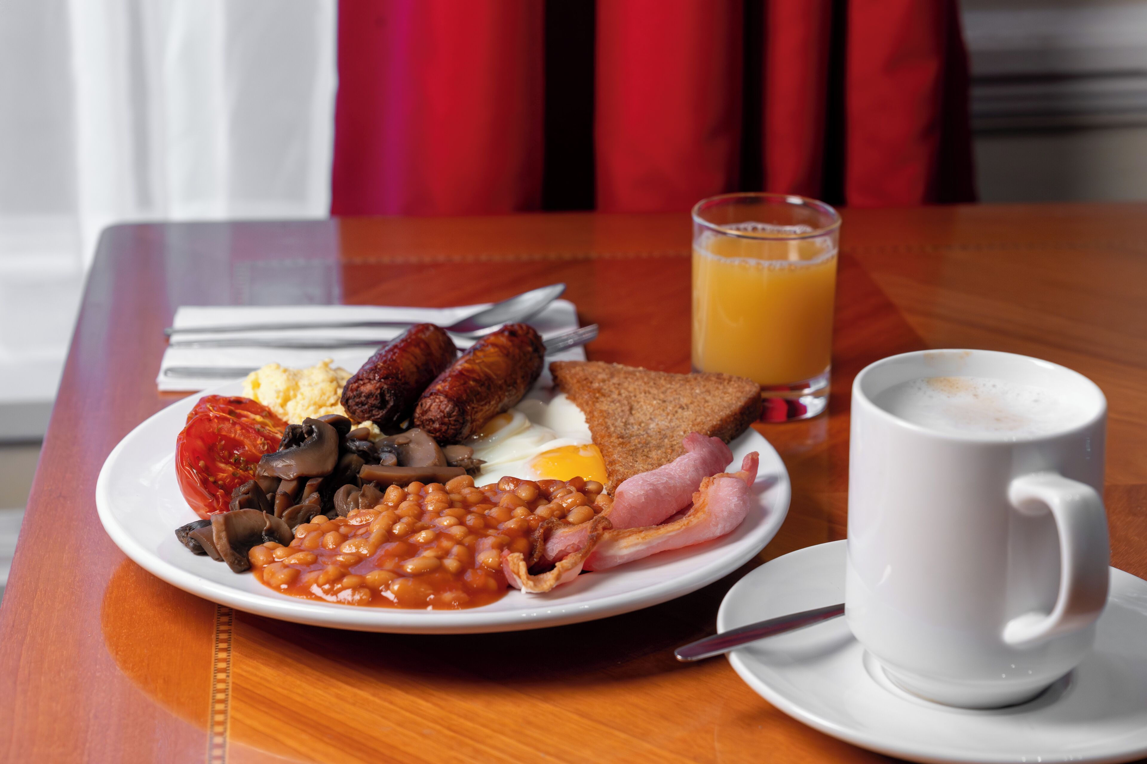 A breakfast plate with beans, sausages, eggs, toast, and ham, accompanied by a glass of orange juice and a cup of coffee on a wooden table.