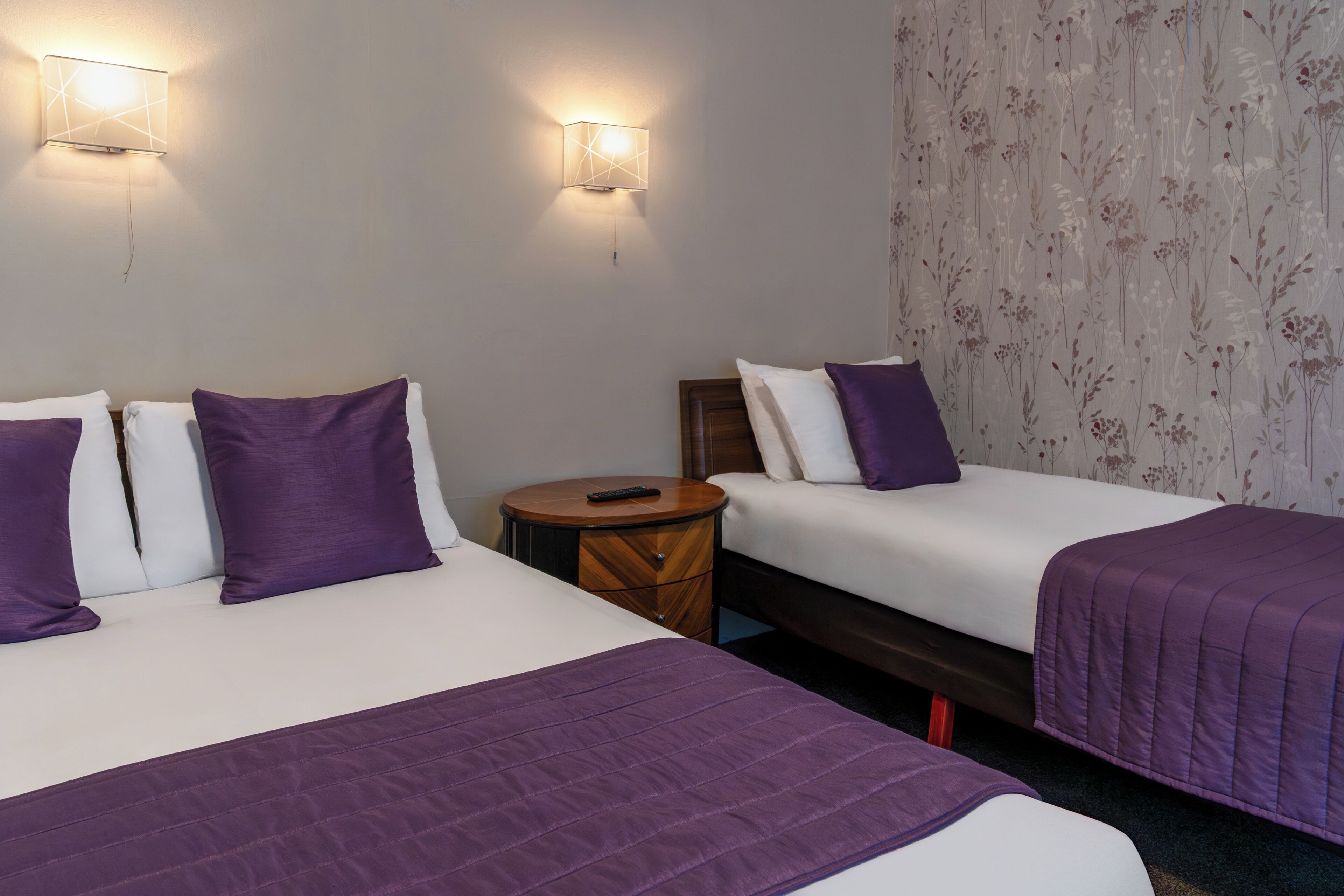 A hotel room with two beds, a round wooden nightstand, and two wall-mounted light fixtures.