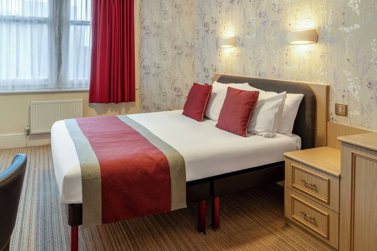 A hotel room with a bed, bedside tables, a chair, a radiator, and a window with curtains.