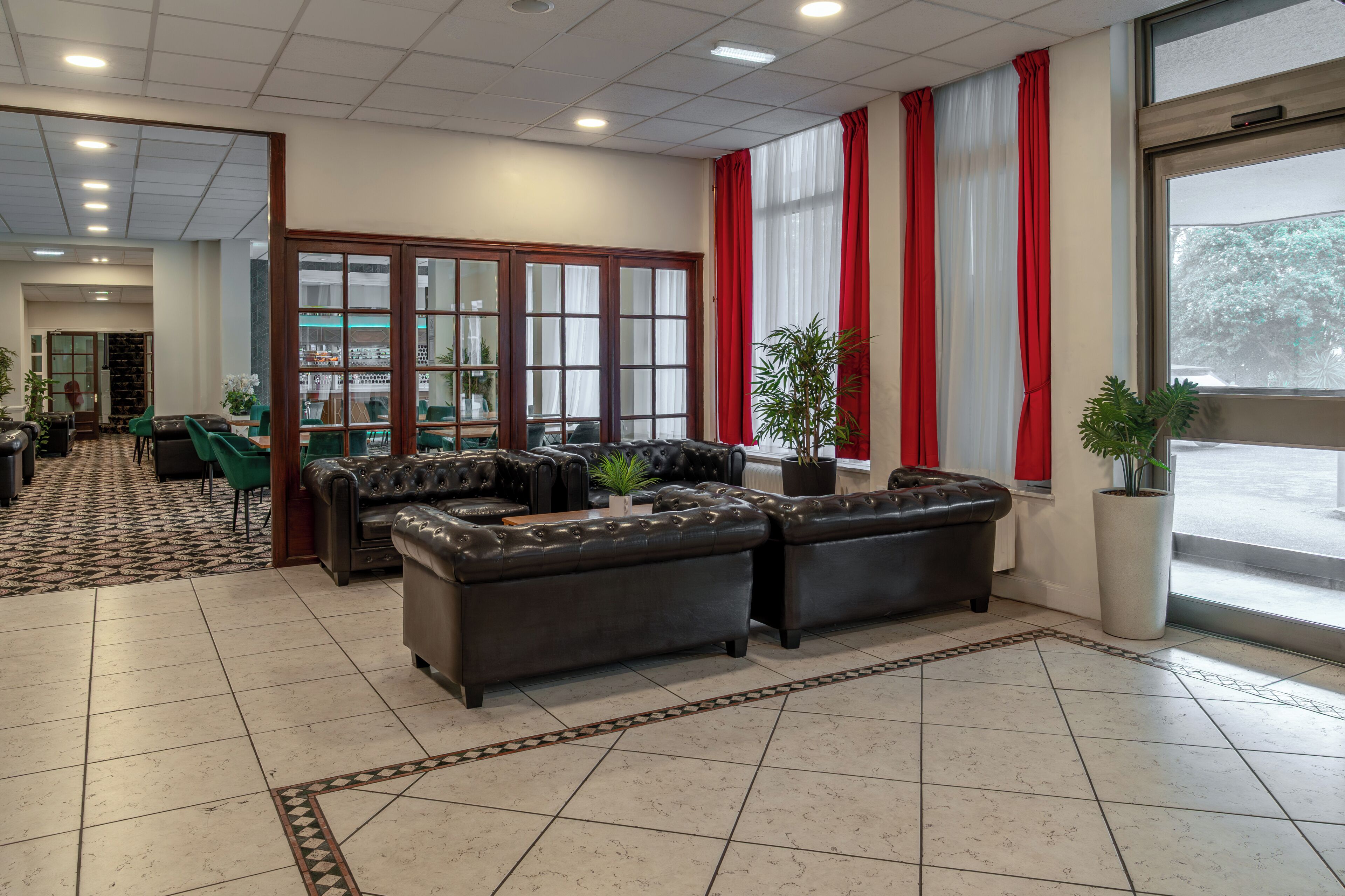 A lobby with leather sofas, a patterned carpet, a large window with red curtains, and a potted plant.