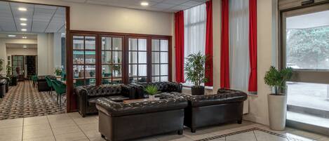A lobby with leather sofas, a patterned carpet, a large window with red curtains, and a potted plant.