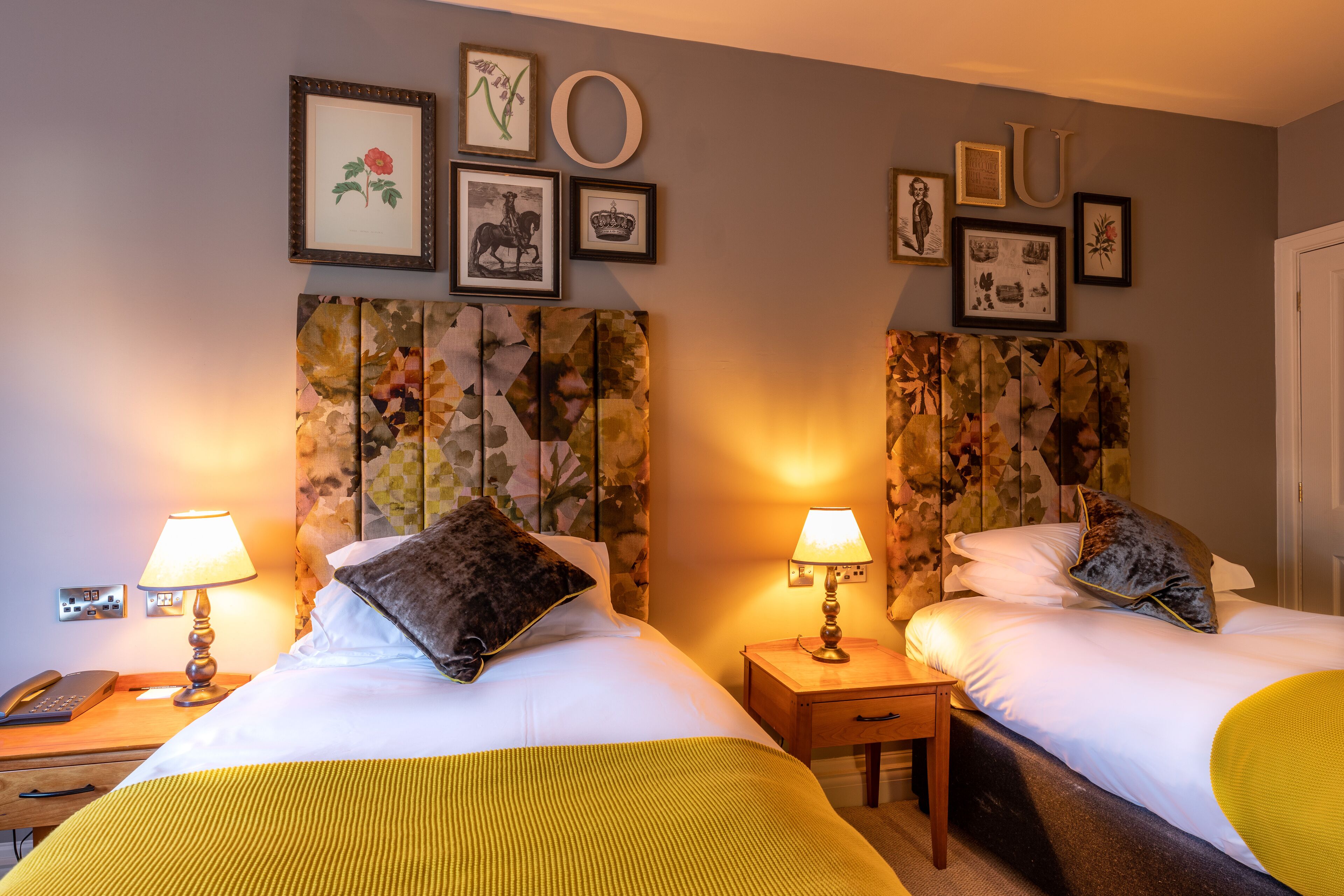 A hotel room with two beds, each with a nightstand, lamps, and framed artwork on the wall.