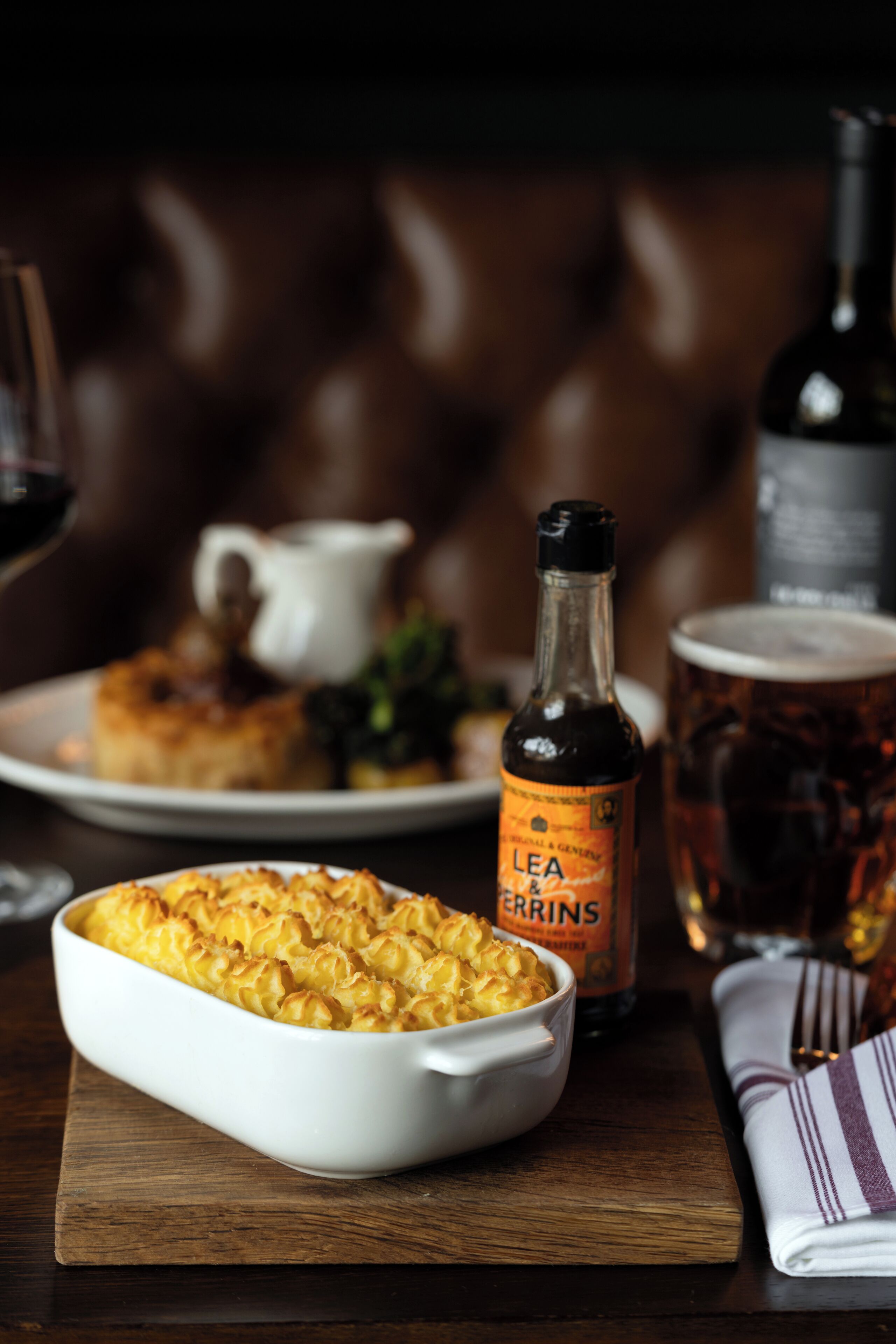 A white ceramic dish of macaroni and cheese, a bottle of Lea & Perrins sauce, and a glass of beer on a wooden table.