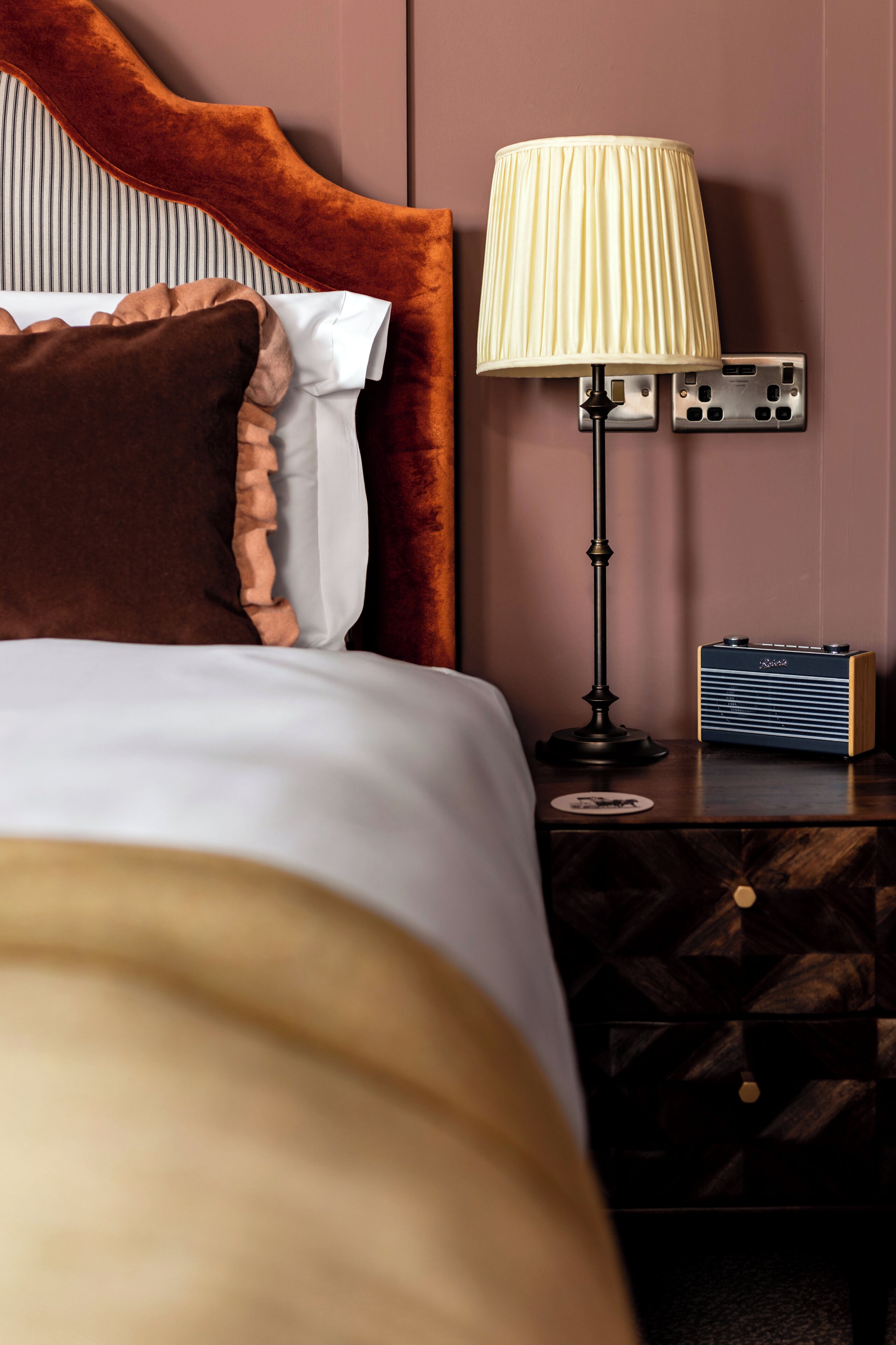 Una cama bien tendida con un cabecero marrón, una lámpara de noche y una radio antigua sobre una mesita de madera.