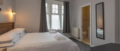A bedroom with a large bed, a bedside table, a chair, a window with curtains, a door leading to a bathroom, and a mirror on the wall.