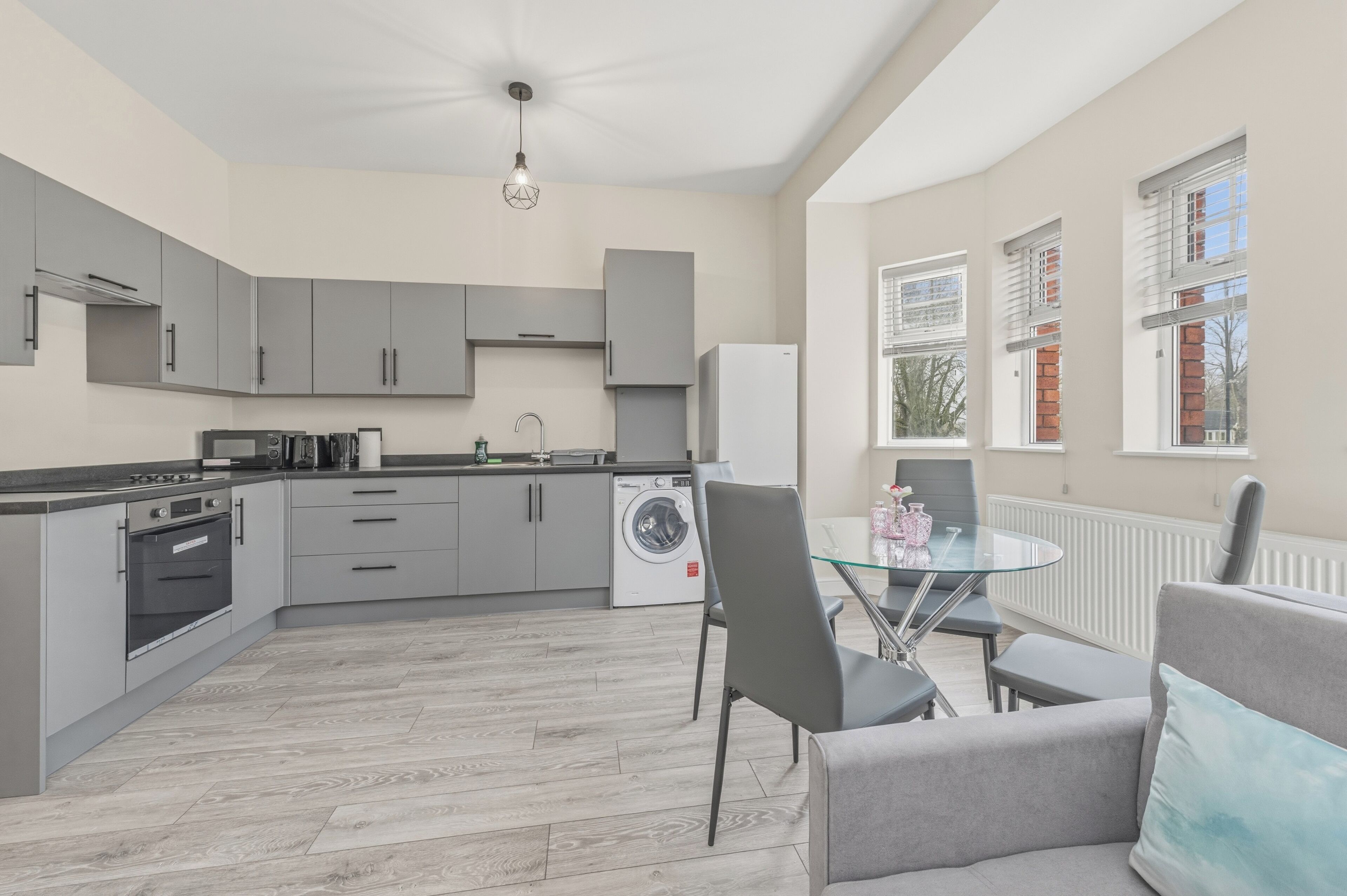A modern kitchen with grey cabinets, a built-in oven, a washing machine, and a dining area with a glass table and chairs.