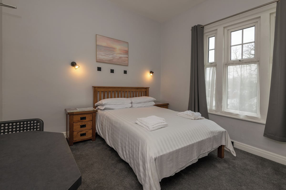 Un dormitorio con cama, mesitas de noche, escritorio y ventana con cortinas.