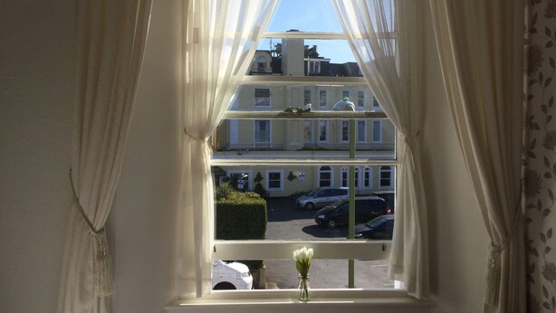 A window with white curtains, a white vase with flowers, and a view of a multi-story building with parked cars. - Newburgh Guesthouse (Torquay)