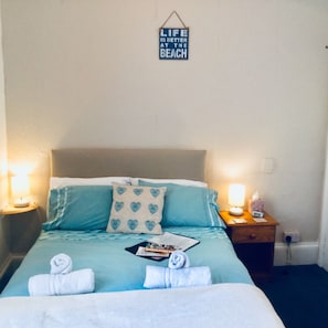 A neatly made bed with a blue and white color scheme, a wall-mounted sign that reads "LIFE IS BETTER AT THE BEACH", and two bedside lamps. - Newburgh Guesthouse (Torquay)