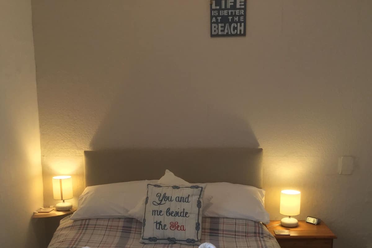 Una cama bien hecha con ropa de cama escocesa, dos almohadas y dos toallas enrolladas. Hay dos lámparas de noche a cada lado de la cama. Un cartel en la pared dice "La vida es mejor en la playa."
