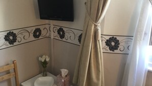 A bathroom with a toilet, a small table with a tissue box and a vase of flowers, a mounted TV, and a curtain. - Newburgh Guesthouse (Torquay)