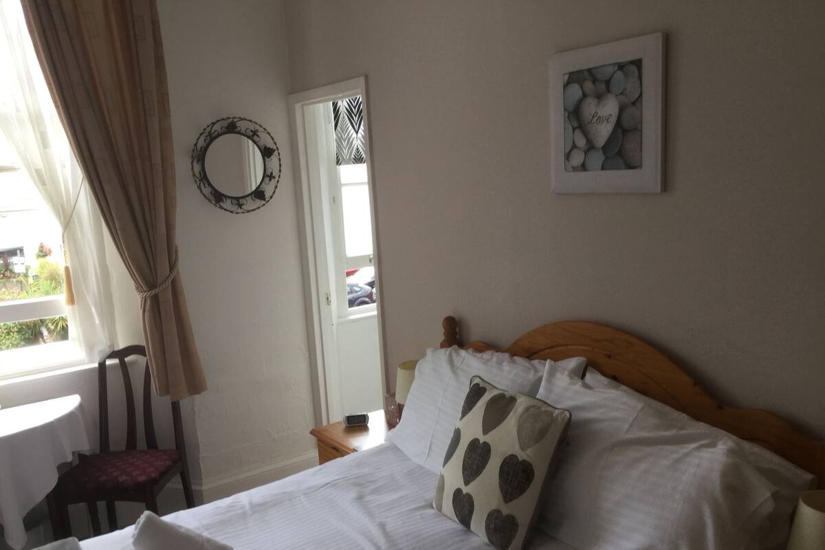 Una habitación de hotel con cama, mesita de noche, silla, ventana con cortinas, espejo y un cuadro en la pared.