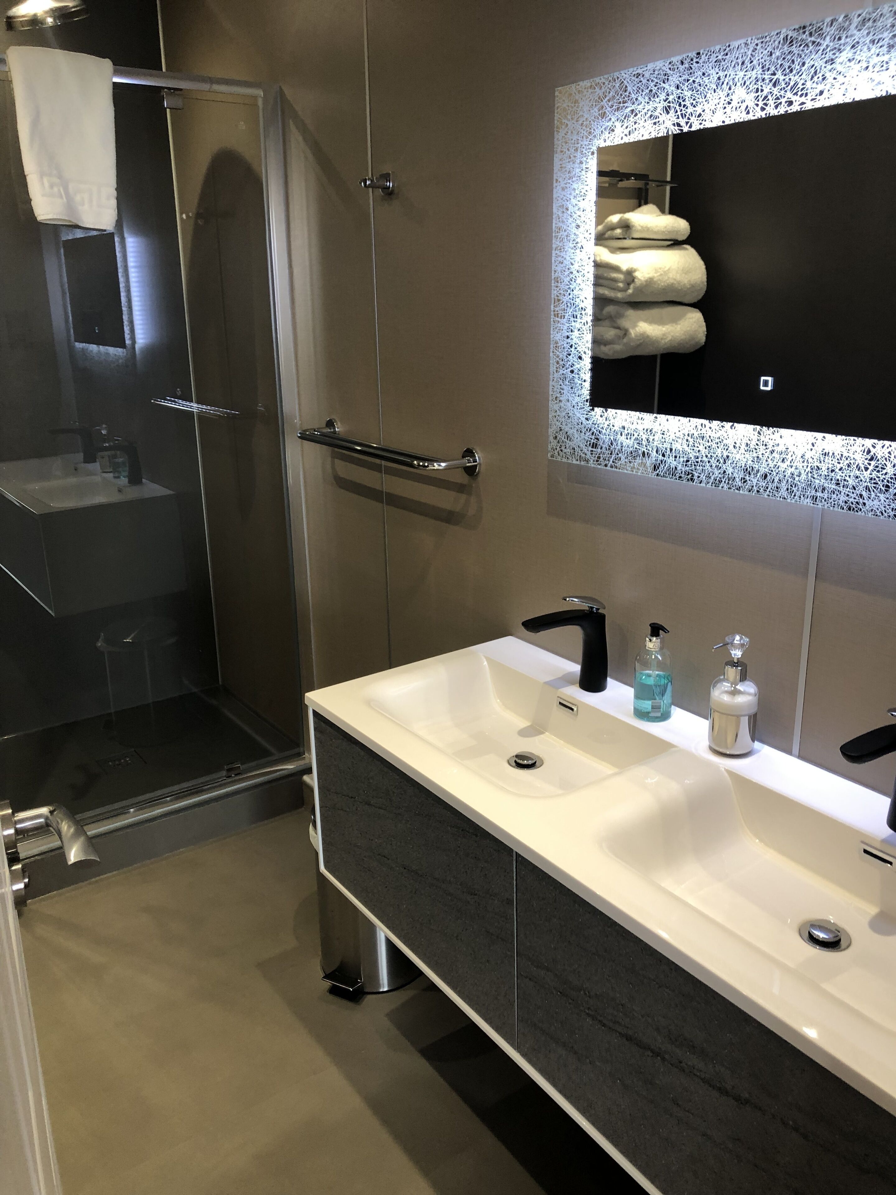 Un baño moderno con cabina de ducha de vidrio, tocador doble y un espejo grande con marco decorativo.