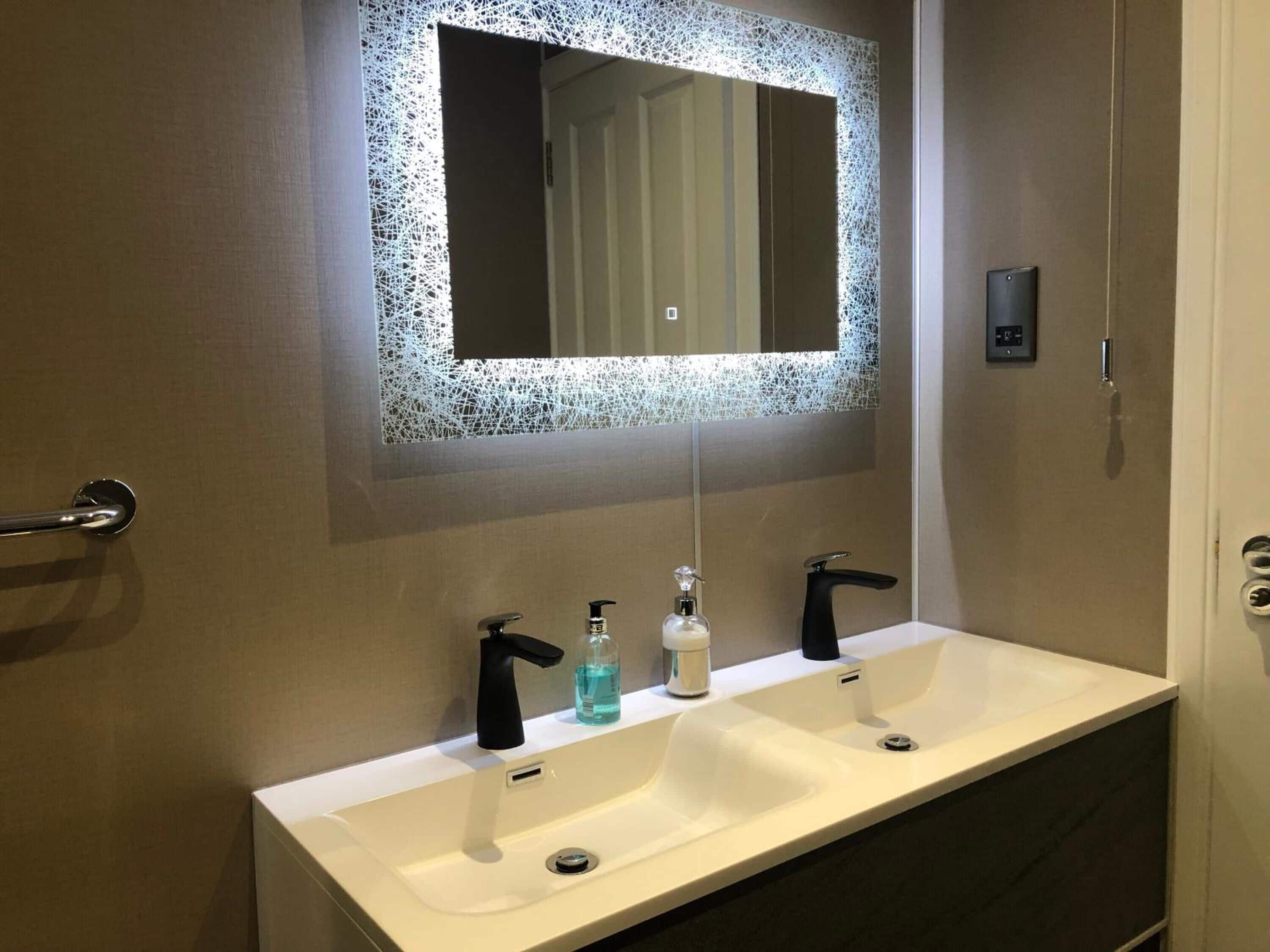 A modern bathroom with a white sink, two black faucets, a soap dispenser, and a mirror with a decorative frame.