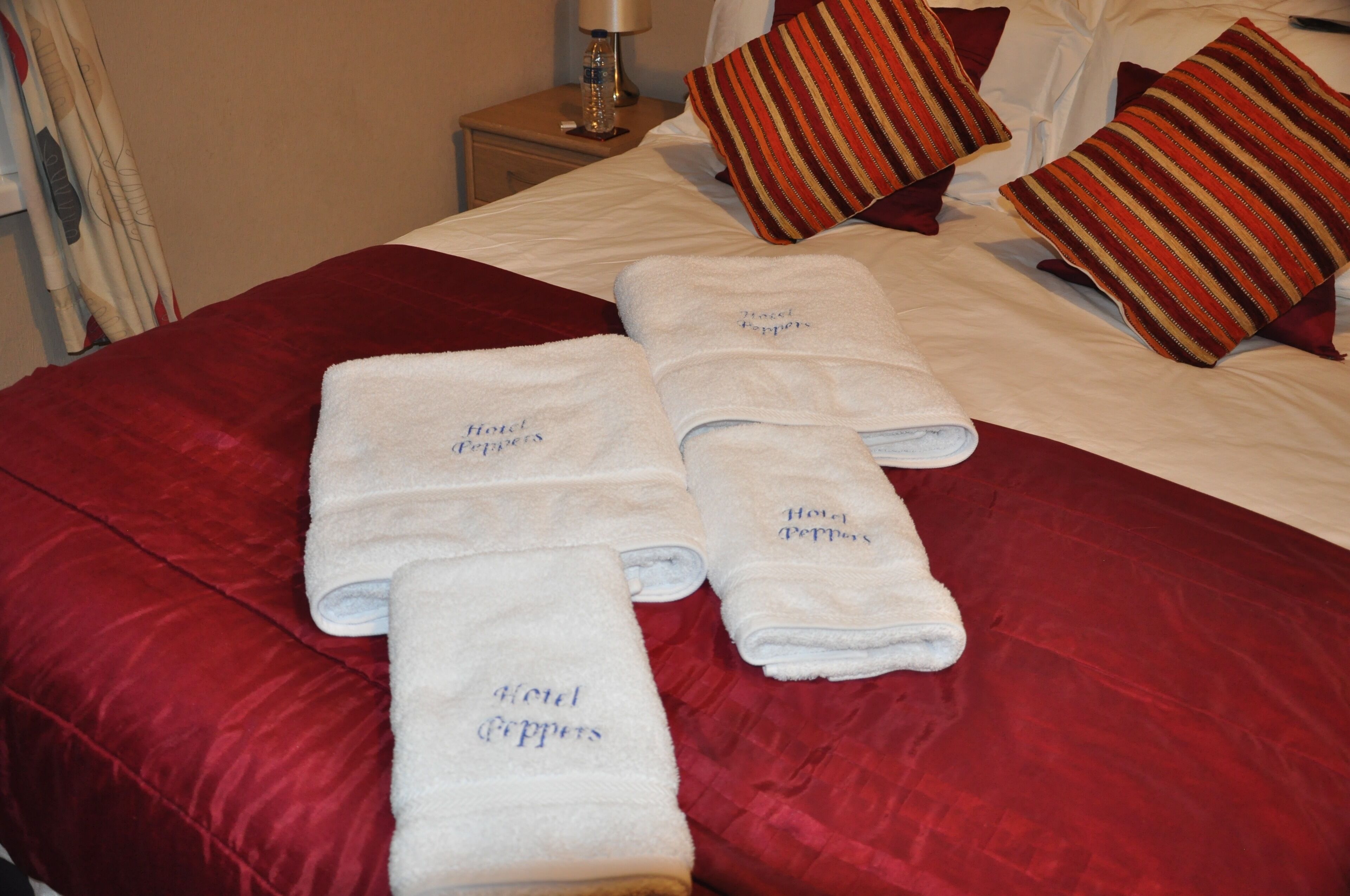 Ein ordentlich bezogenes Bett mit roter Bettwäsche und vier weißen Handtüchern mit dem Schriftzug 'Hotel Peppermint', einem gestreiften Kopfkissen und einer Lampe auf einem Nachttisch.