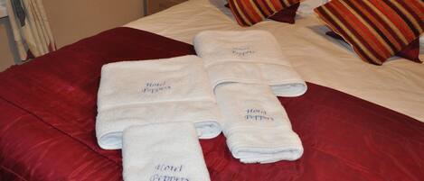 Una cama bien hecha con ropa de cama roja y cuatro toallas blancas con el texto 'Hotel Peppermint', un almohadón a rayas y una lámpara en una mesita de noche.