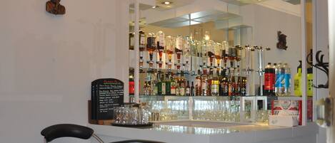 Zona de bar con pared espejada, taburetes y una variedad de botellas y vasos.
