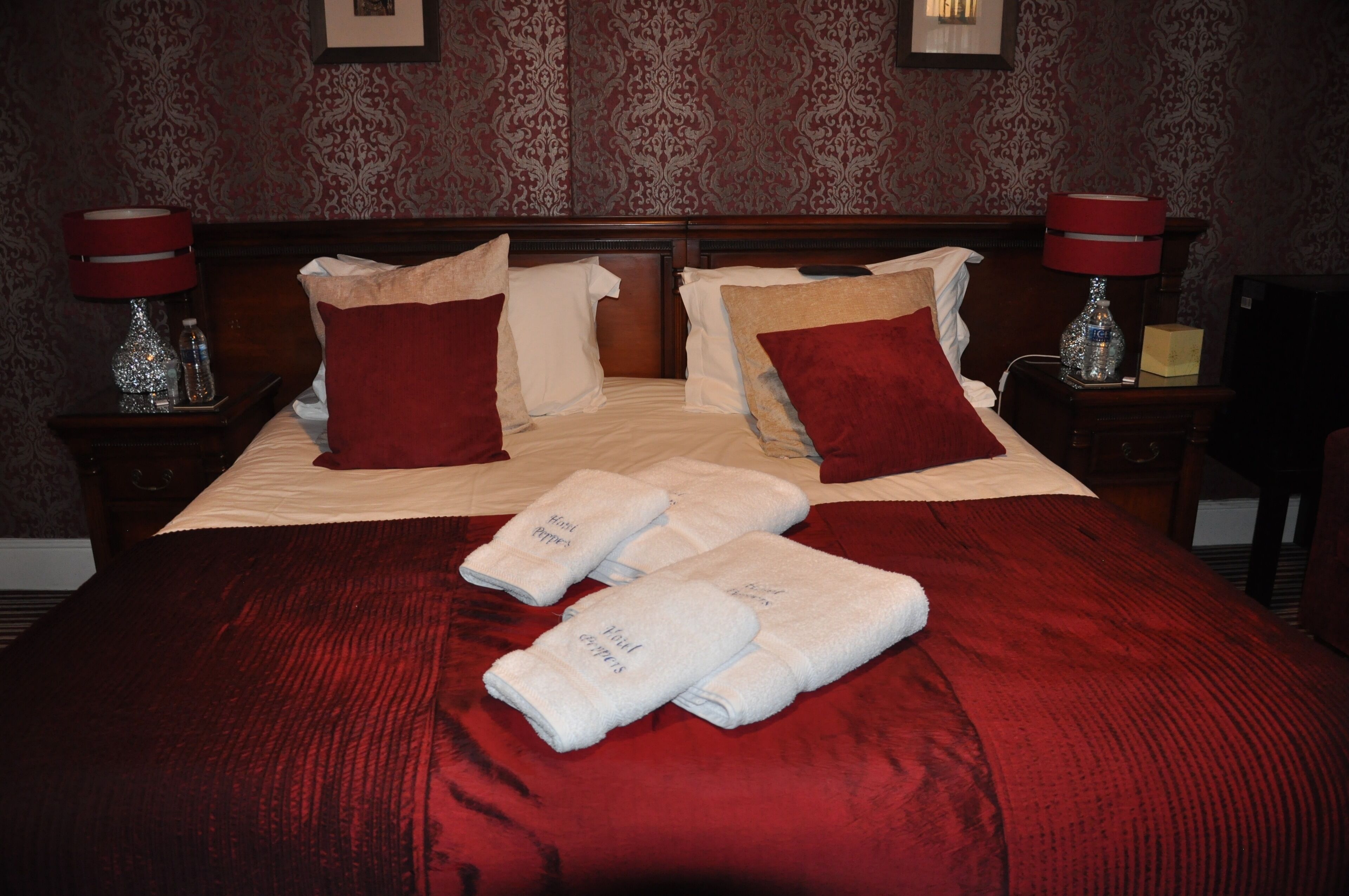 A neatly made bed with red bedding, white pillows, and a red headboard. There are two bedside tables with lamps and a small bottle on one.
