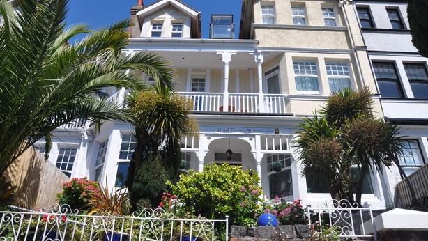 A multi-story hotel with a white facade, balconies, and a garden with various plants and flowers. - Hotel Peppers (Torquay)