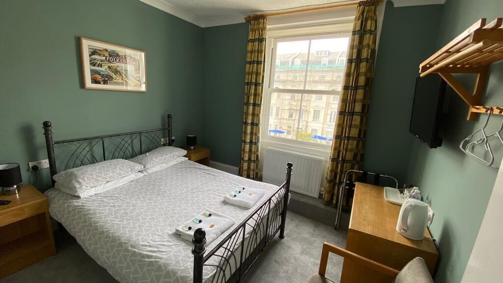 A bedroom with a bed, bedside tables, a desk with a chair, a window with curtains, and a wall-mounted TV.