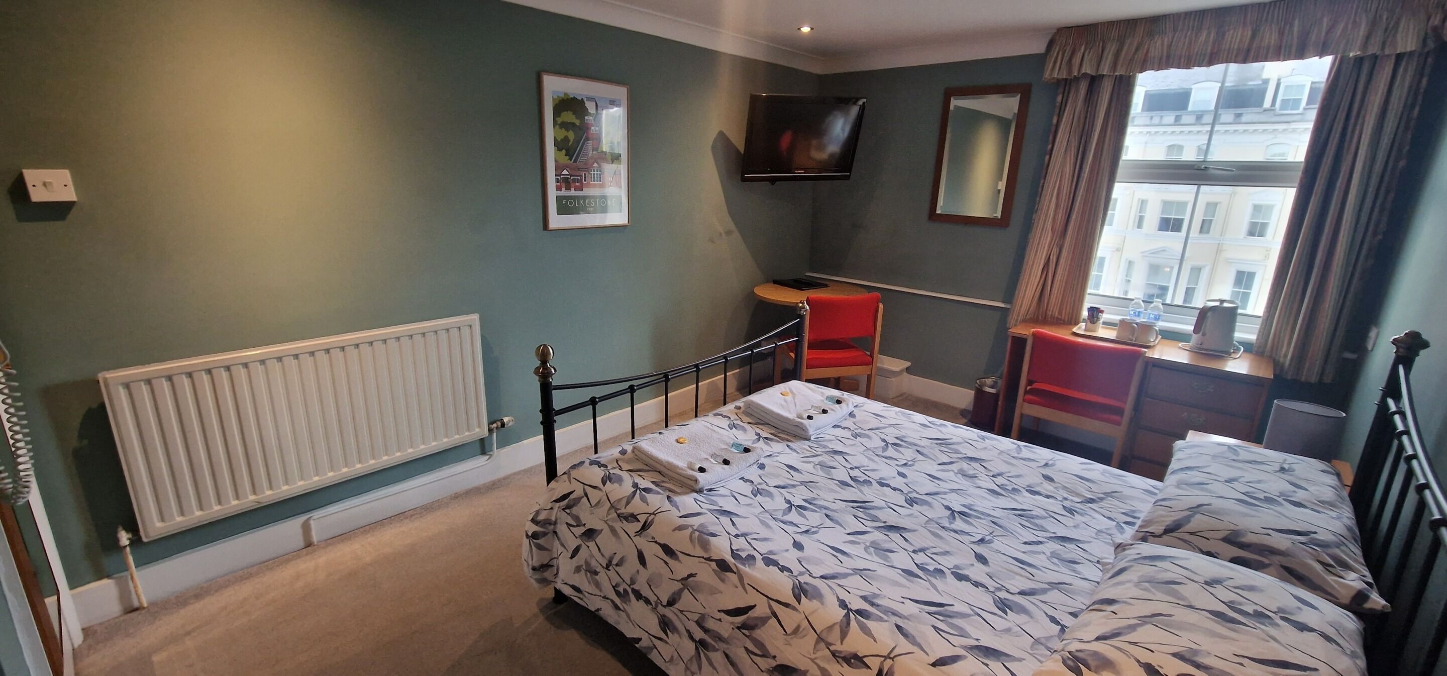 A bedroom with a bed, a desk, a chair, a radiator, a TV, and a window with curtains.