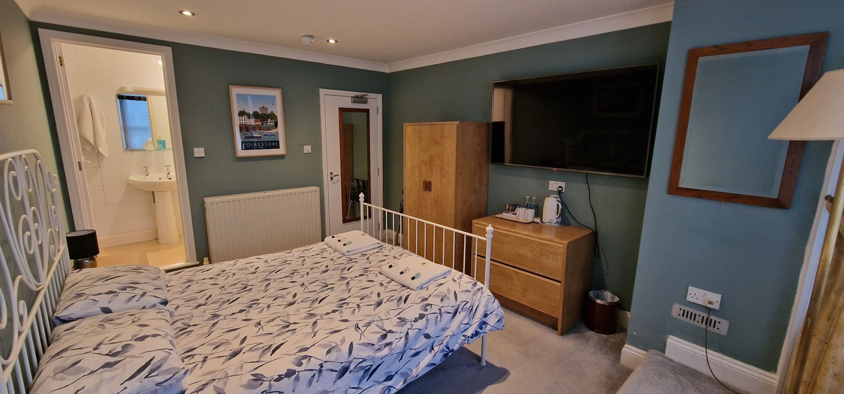 Un dormitorio con cama, armario de madera, mesita de noche, espejo, televisor y baño visible a través de una puerta abierta.