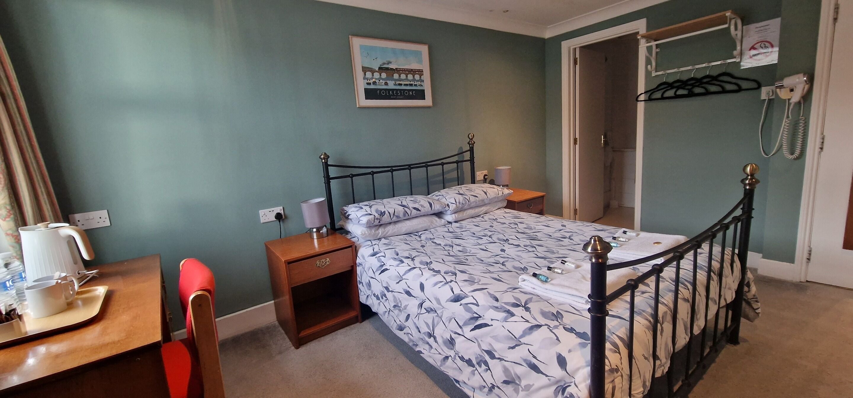 A bedroom with a bed, bedside table, and a chair. There is a wall-mounted telephone and a framed picture above the bed.