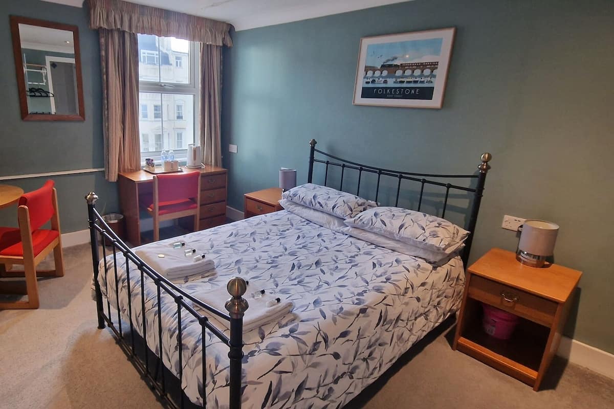 A bedroom with a bed, a desk, a chair, a TV, and a painting on the wall.