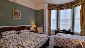 Quarto de hotel com duas camas, uma escrivaninha, uma cadeira, um radiador e uma janela com cortinas.