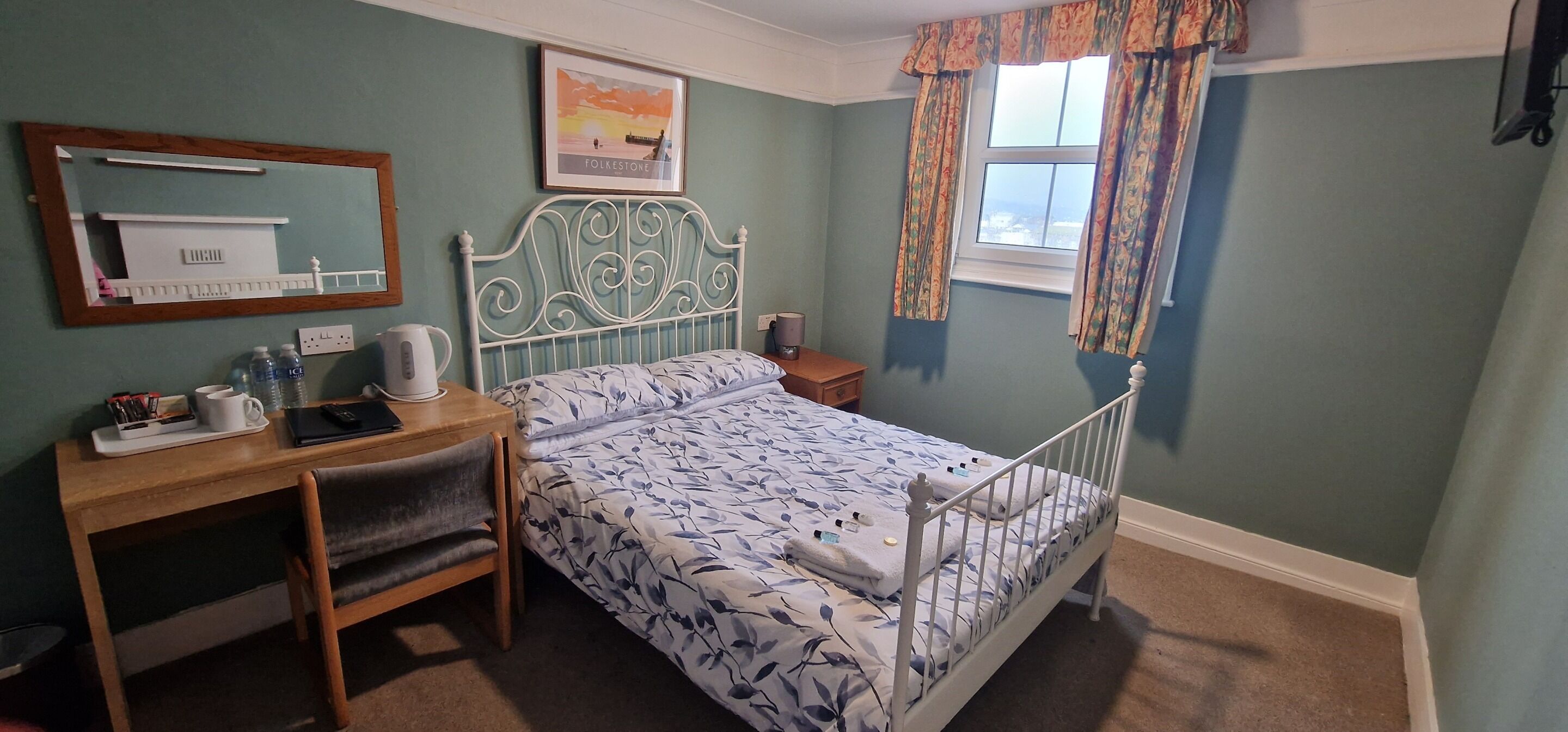 Un dormitorio con cama, escritorio, silla, espejo y ventana con cortinas.