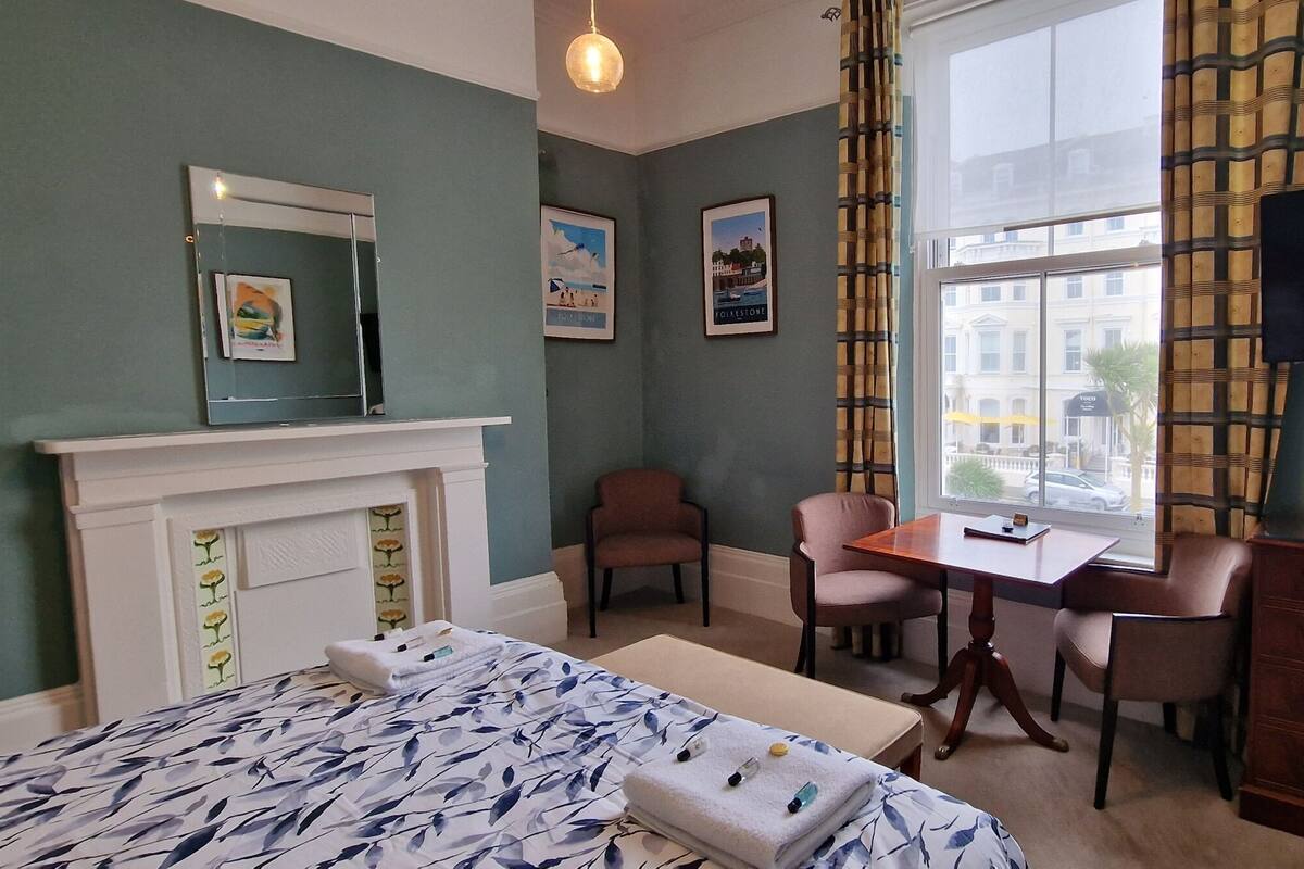 A hotel room with a bed, a desk, two chairs, a dresser, a TV, and a window with a view of buildings.