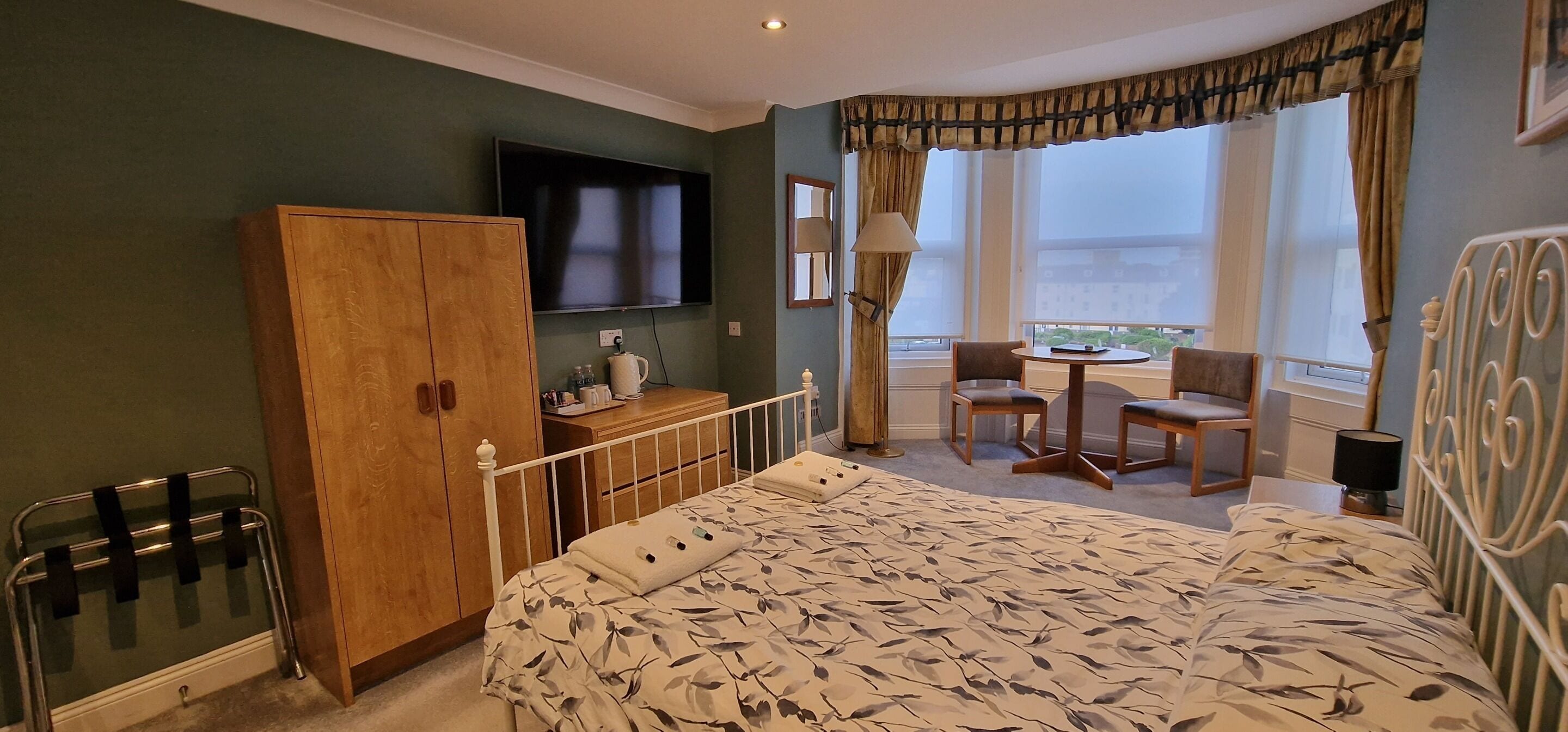 A hotel room with a bed, a wooden wardrobe, a television, a small table with chairs, and a window with curtains.