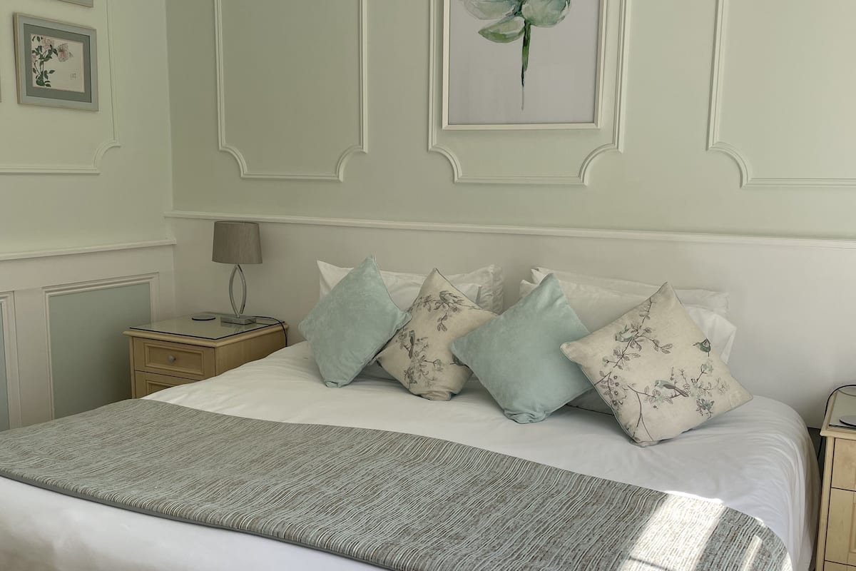 A neatly made bed with a grey blanket and floral pillows, a bedside table with a lamp, and framed artwork on the wall.
