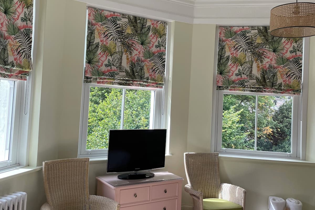 A bedroom with a television on a dresser, two wicker chairs, a bed with a pink blanket, and windows with tropical leaf-patterned curtains.