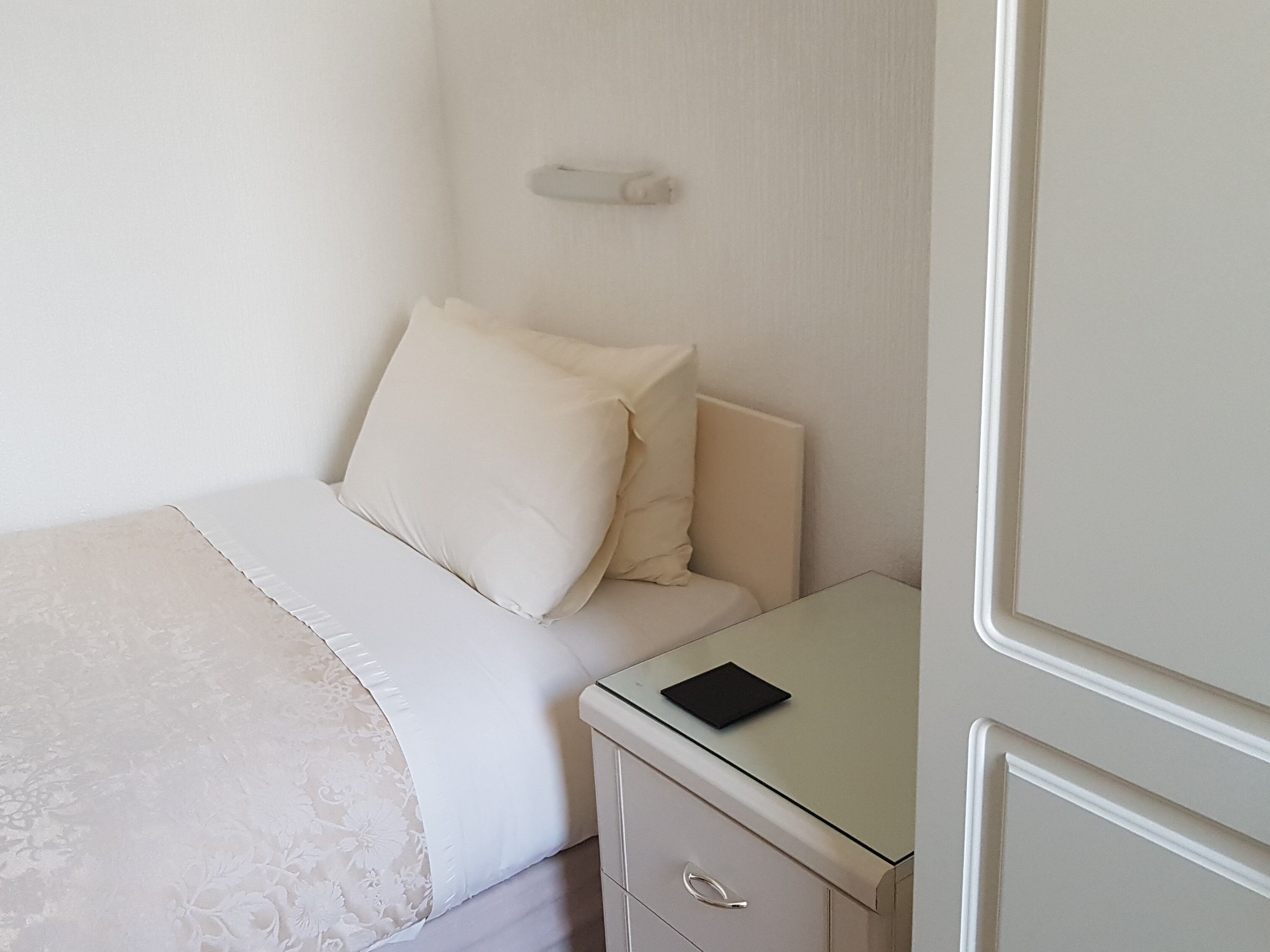 Una cama bien hecha con edredón blanco y almohadas, una mesita de noche con un objeto negro encima y un cajón con asa plateada.