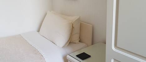 Una cama bien hecha con edredón blanco y almohadas, una mesita de noche con un objeto negro encima y un cajón con asa plateada.