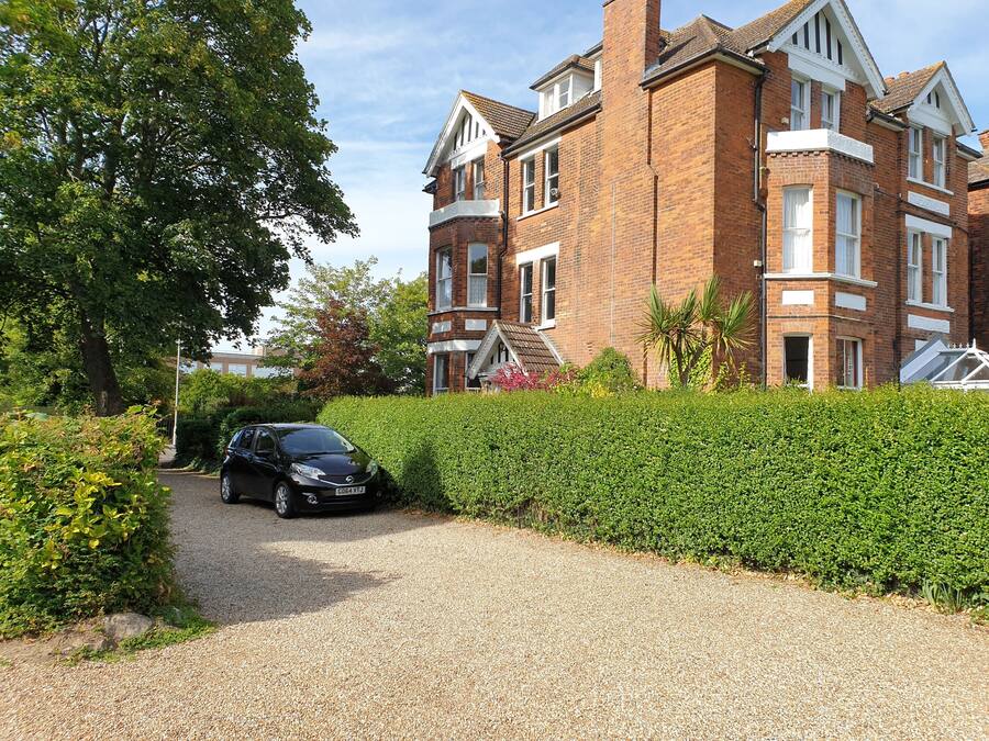 A residential building with a brick facade, multiple windows, and a driveway leading to a parked car.