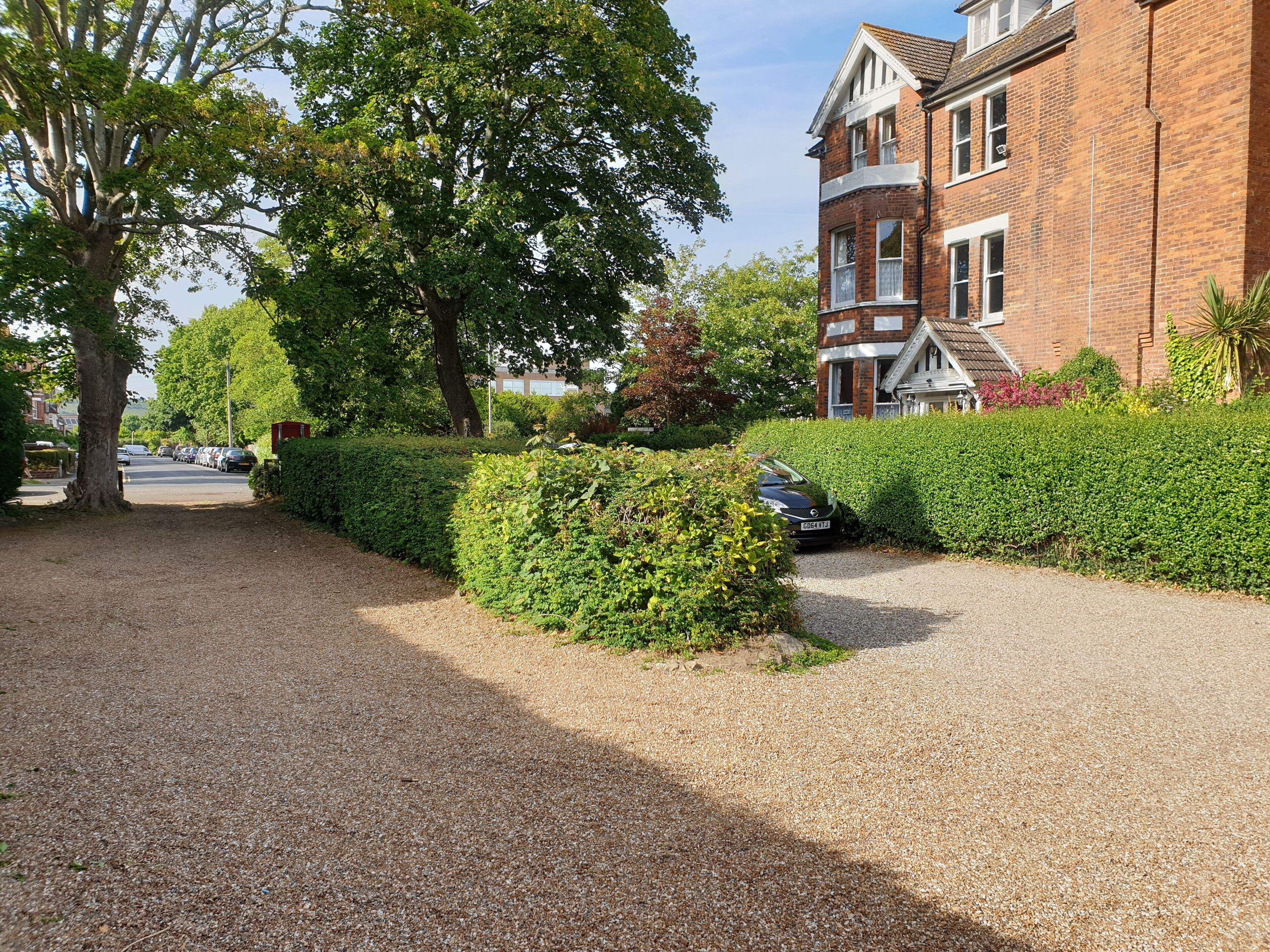 A residential street with a gravel driveway, a parked car, and a brick house with a white window frame.