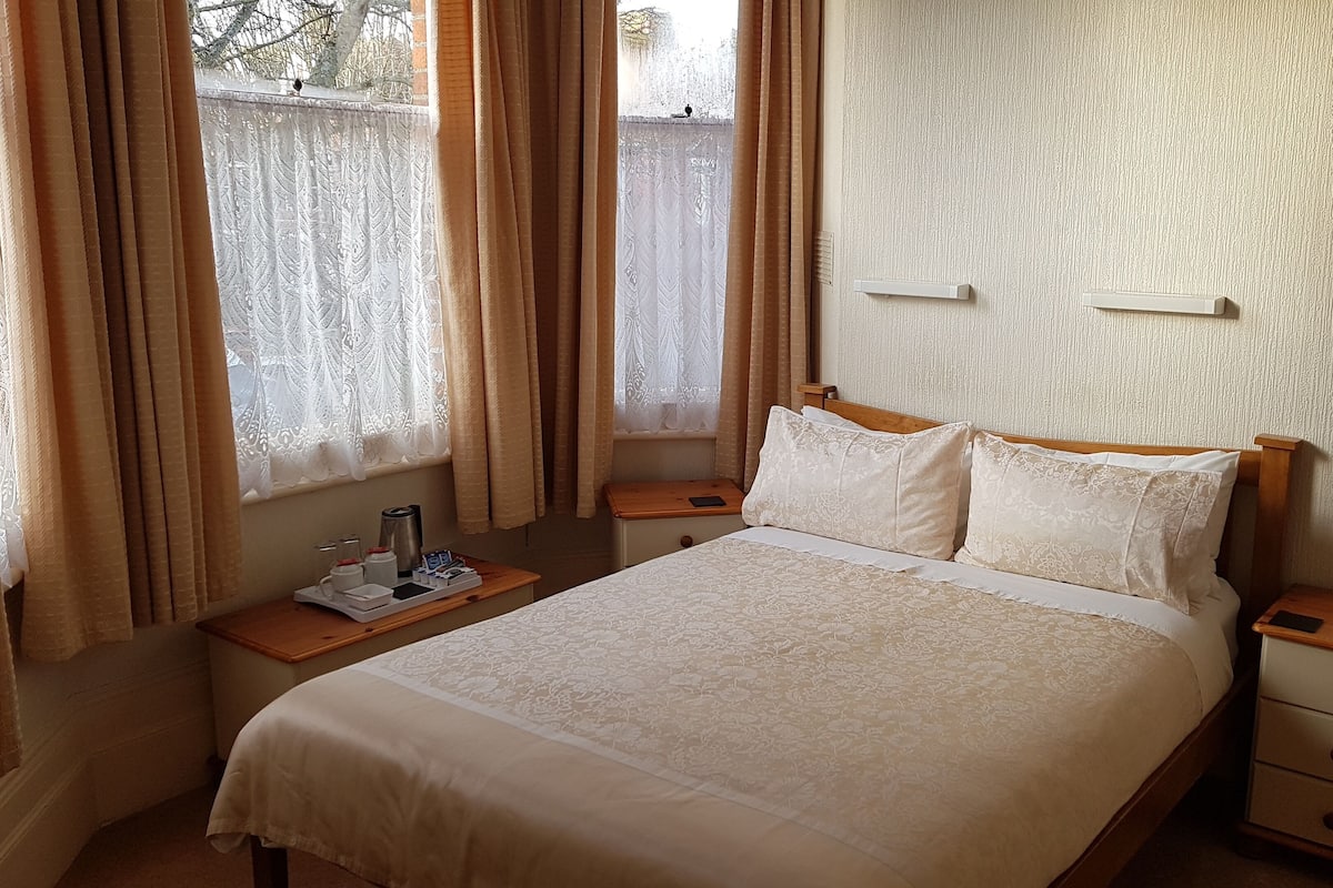 Een opgemaakt bed met een bloemenprintdeken, twee nachtkastjes met lampen, een raam met dunne gordijnen en een structuurwand.