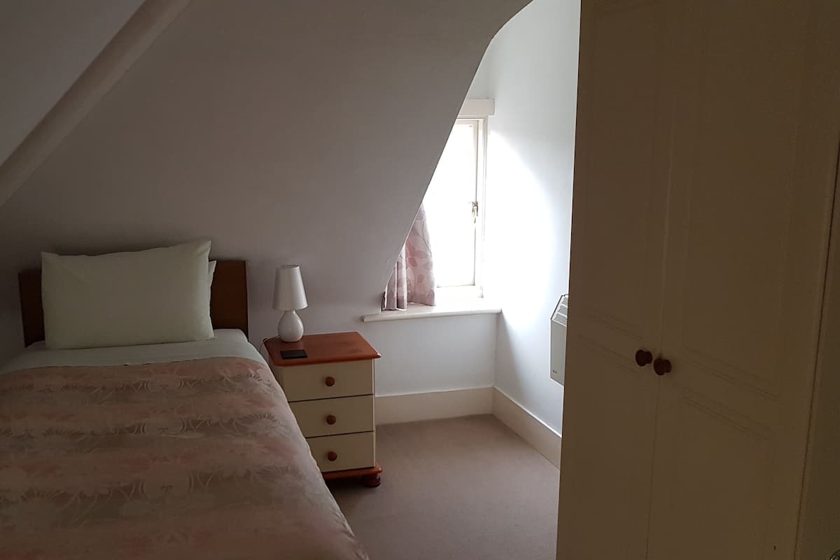 Een slaapkamer met een bed, nachtkastje, lamp, raam met gordijnen en een deur.