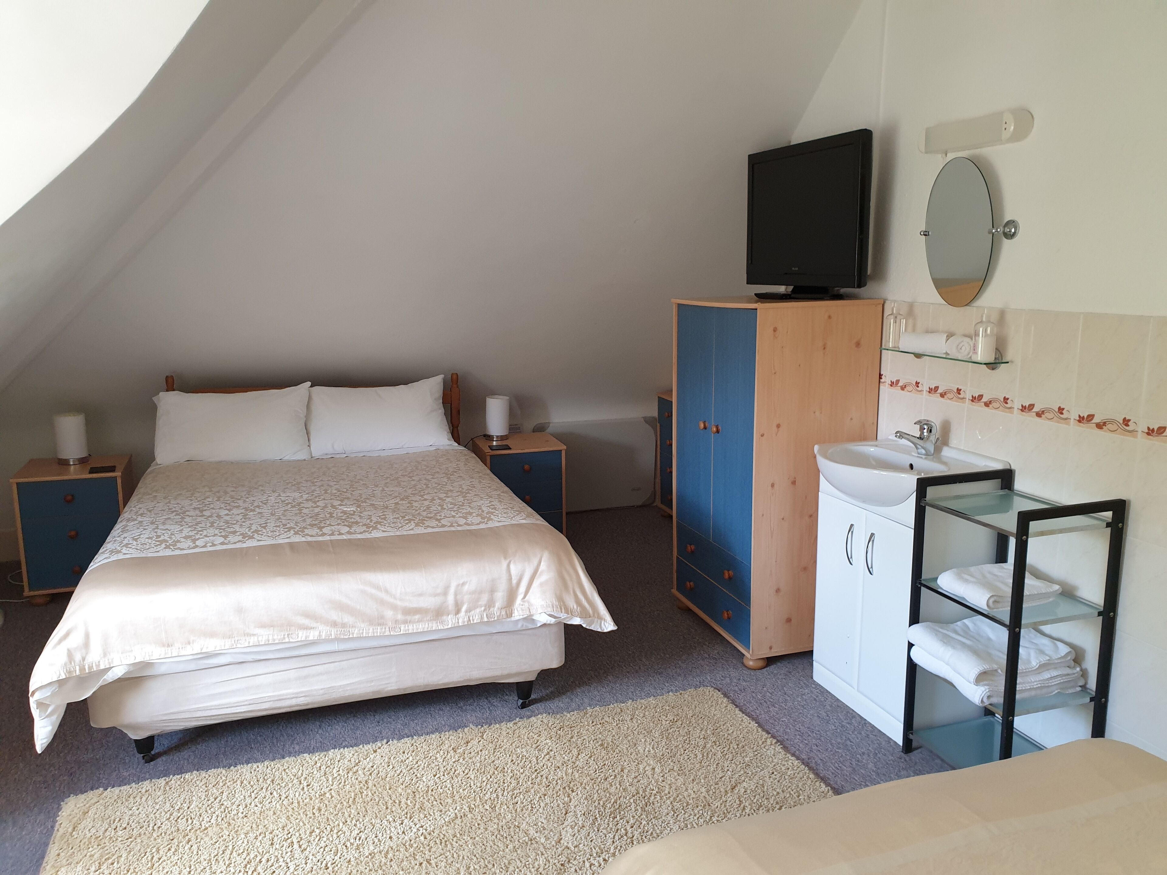 A bedroom with a bed, bedside tables, a TV, a wardrobe, a mirror, and a bathroom with a sink and towel rack.