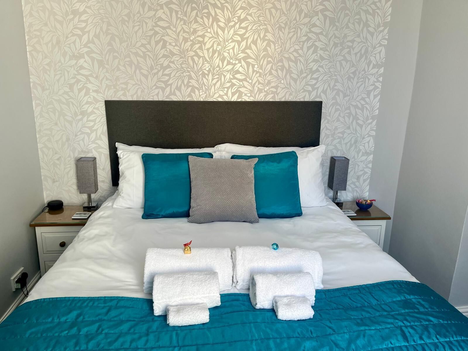 Una cama bien hecha con ropa de cama blanca y aguamarina, dos mesitas de noche con lámparas, y un estampado floral en el papel tapiz de la pared.