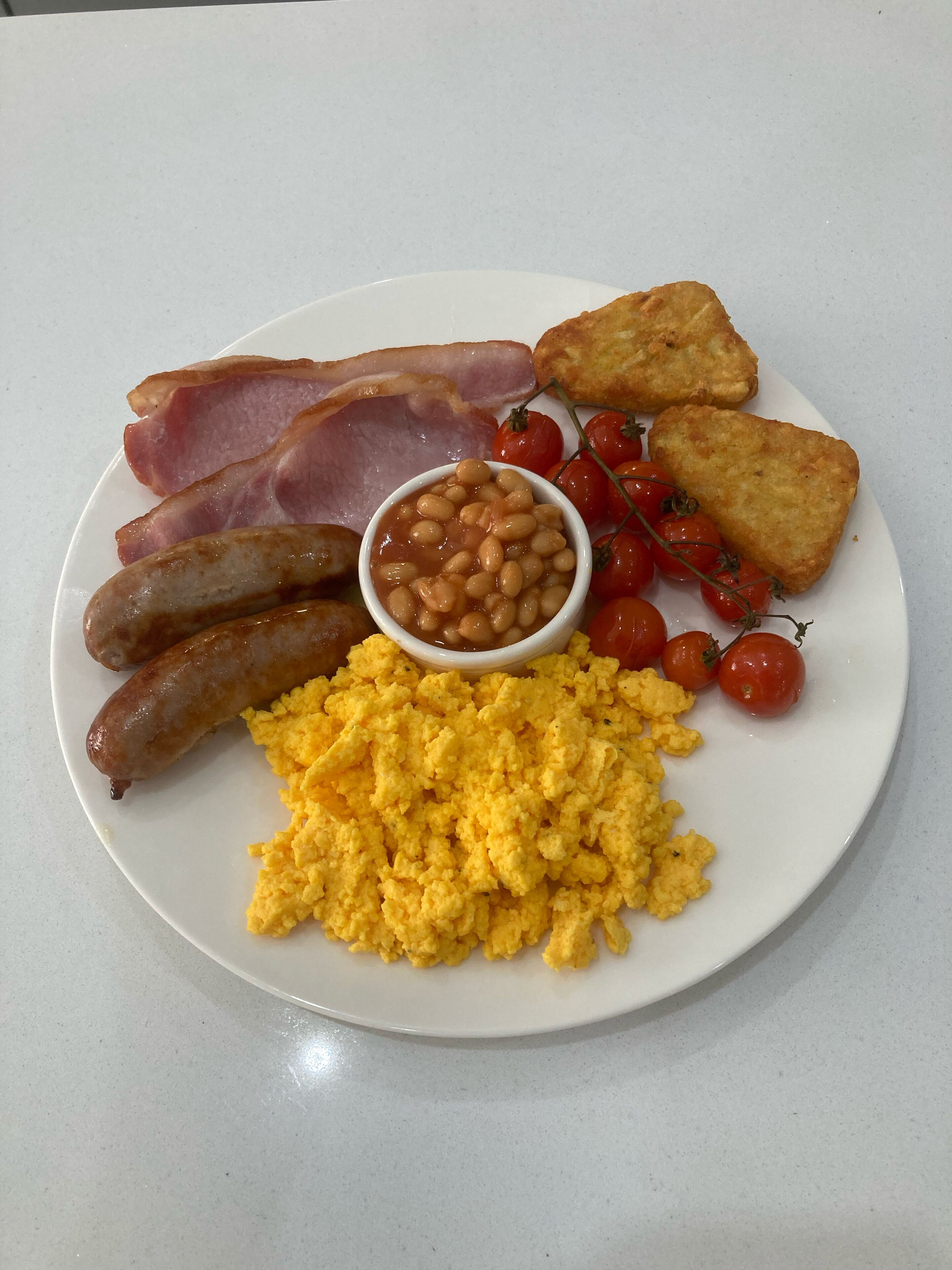 A plate of breakfast food including sausages, bacon, scrambled eggs, beans, tomatoes, and toast.