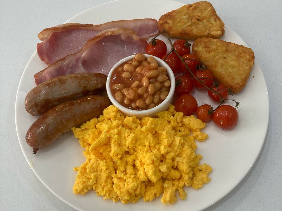 A plate of breakfast food including sausages, bacon, scrambled eggs, beans, tomatoes, and toast.